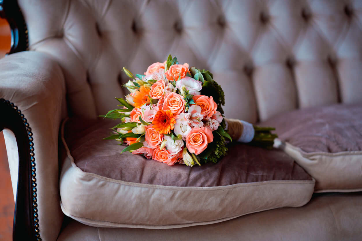 Brautstrauß "Abiona" in kräftigem Korallfarbton mit Rosen, Gerbera und Alstromeria auf einem Sofa.